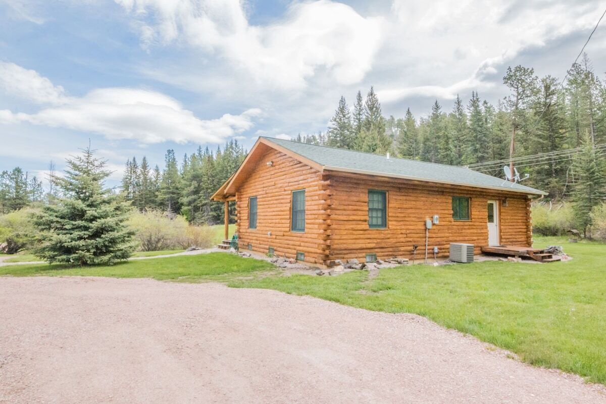 A log cabin with a gravel driveway and a forested backdrop.