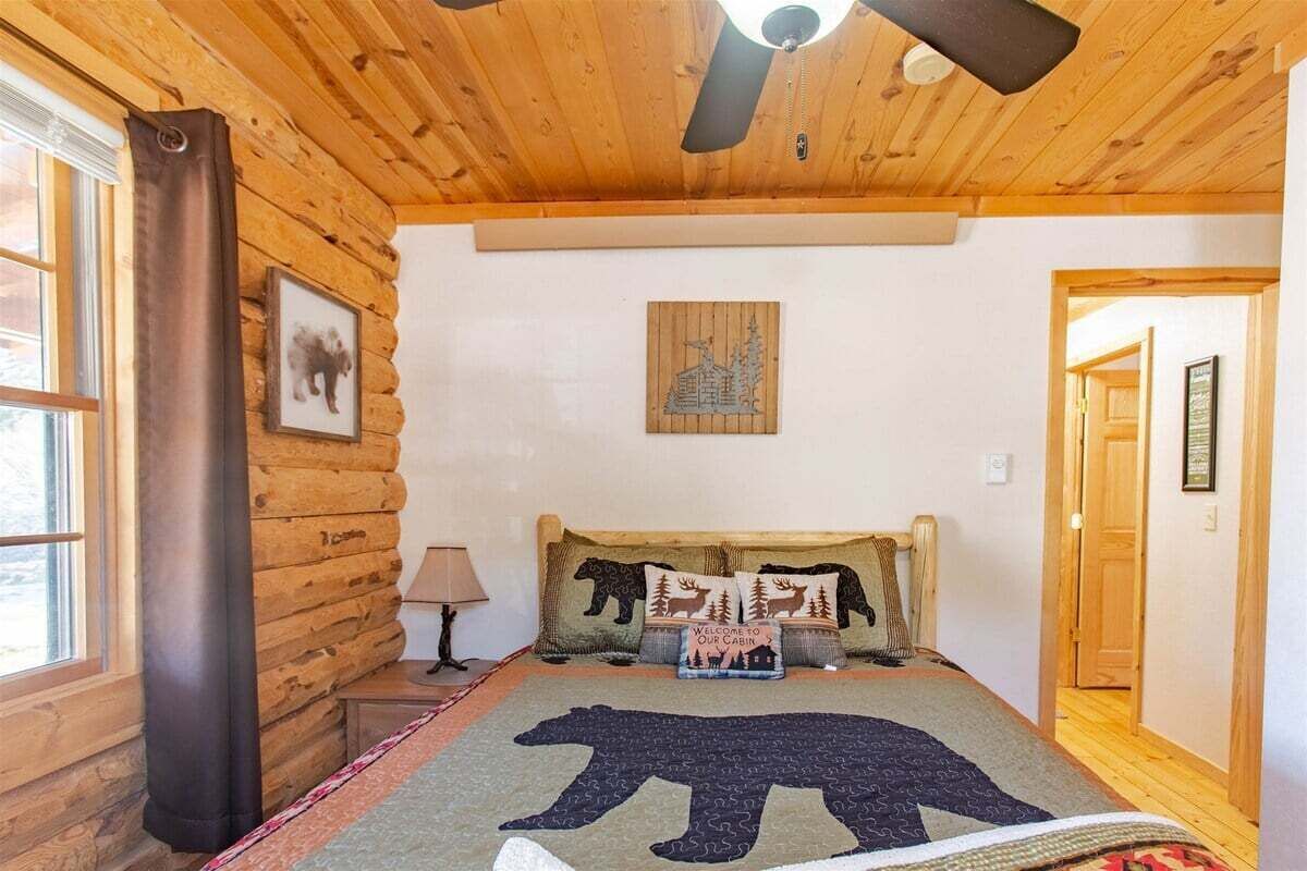 Une chambre à coucher de style chalet, avec un lit, un ventilateur de plafond, une fenêtre avec des rideaux et une porte.