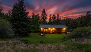 Une cabane en rondins avec un foyer et des sièges d’extérieur, entourée d’arbres et baignée par la lumière d’un coucher de soleil.