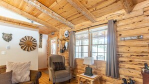 Interieur van een hut met houten wanden, een open haard, een bank, een stoel, een lamp en een bordje met wandelroutes.