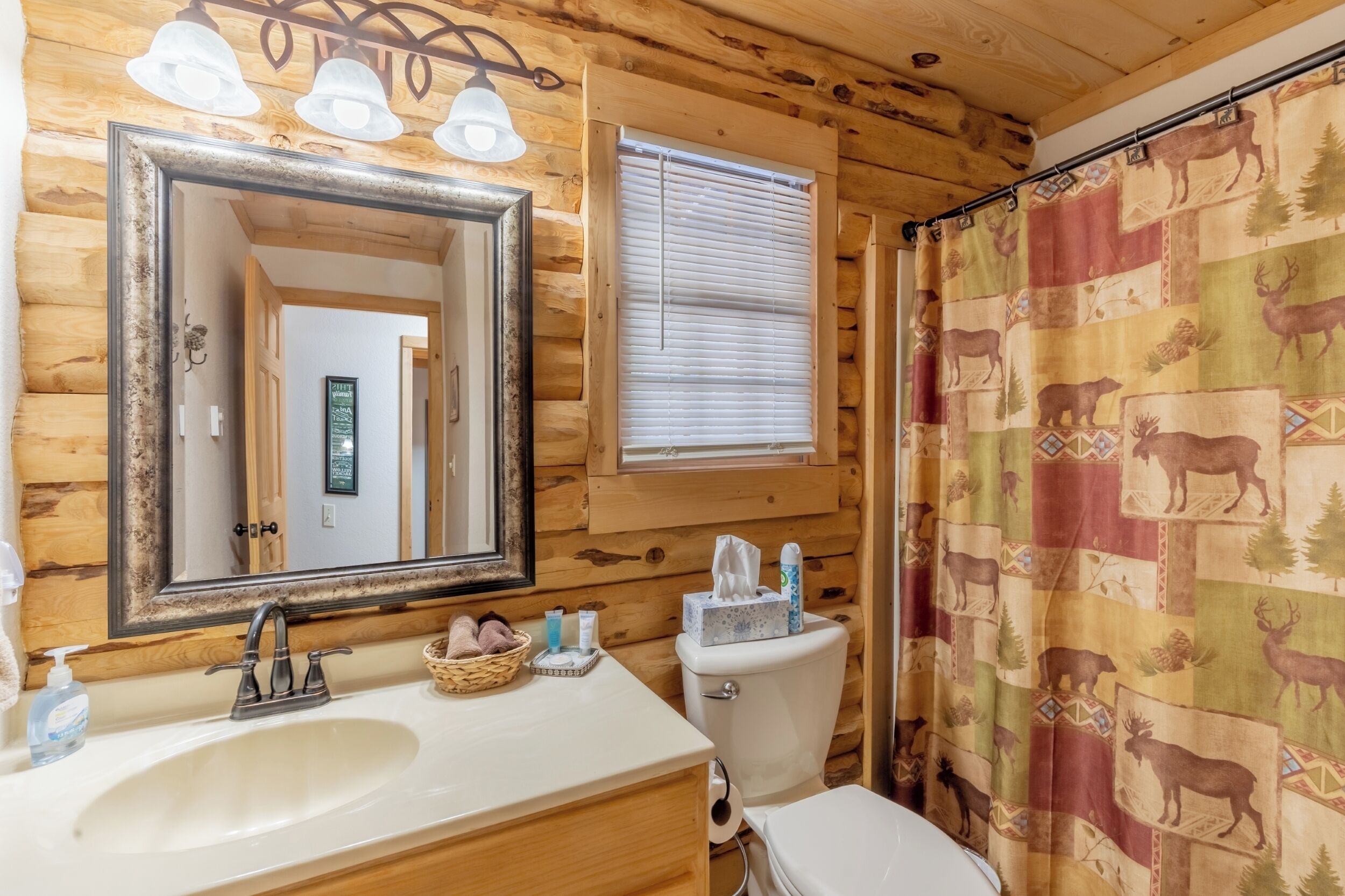 Baño de cabaña de madera con lavamanos, espejo, inodoro y cortina de ducha con estampados de animales.