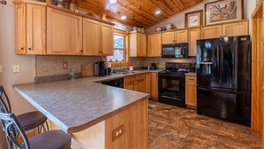 Una cocina con armarios de madera, encimera de granito, refrigerador negro y suelo de baldosas de piedra.