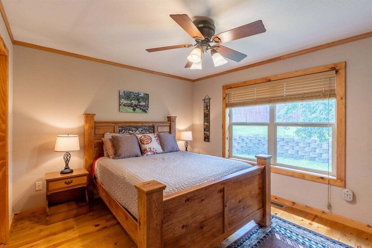 Une chambre à coucher avec un lit en bois, une table de chevet, un ventilateur de plafond, une fenêtre avec des stores et un petit tapis.