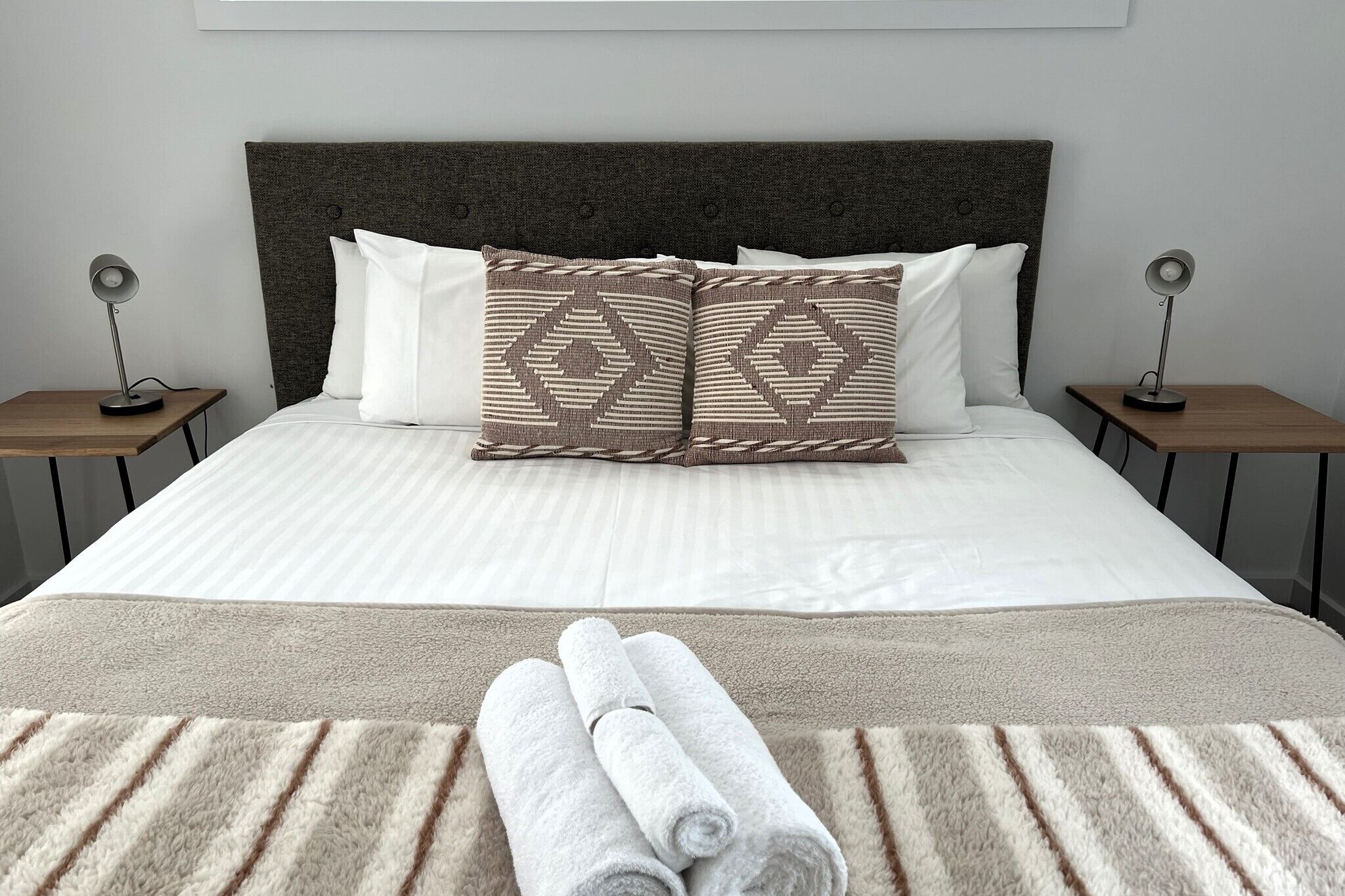 A neatly made bed with a striped white and beige duvet, two decorative pillows with a geometric pattern, and two rolled towels on a beige rug.