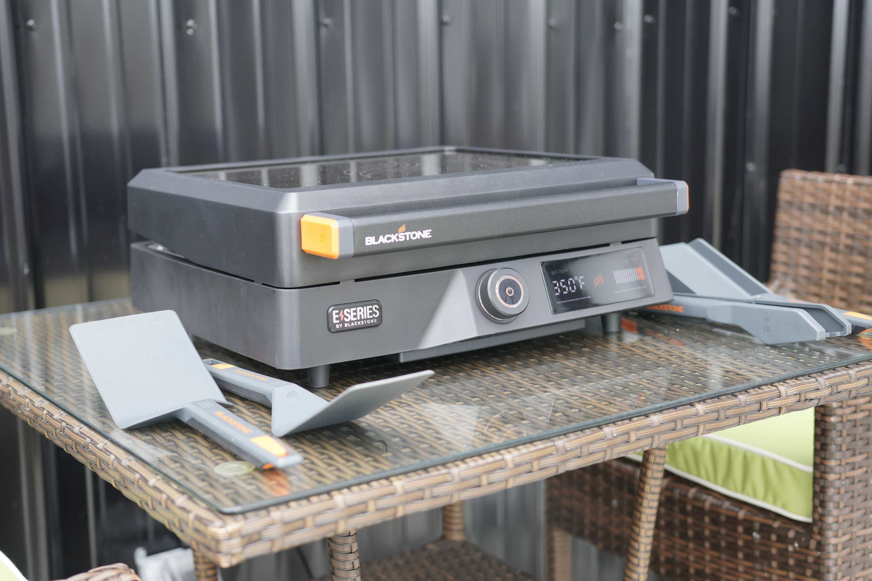A portable grill with a control panel and a small display, placed on a glass table outdoors.
