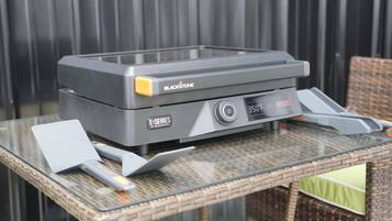A portable grill with a control panel and a small display, placed on a glass table outdoors.