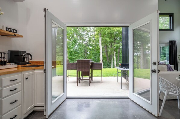 A room with a kitchen area, a dining table with chairs, and a view of a green outdoor space.