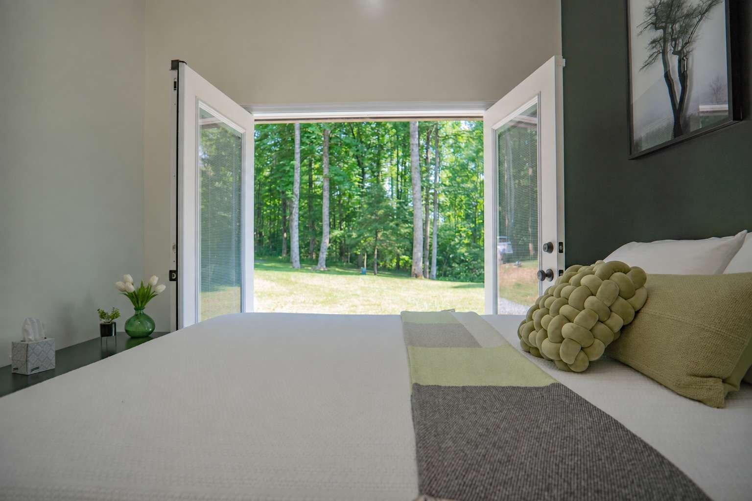 A bedroom with a large bed, a view of a green landscape through open doors, and a framed artwork on the wall.