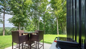 Un área de comedor al aire libre con mobiliario de mimbre y una parrilla, rodeada por un paisaje verde exuberante y árboles.
