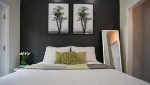 Un dormitorio con una cama, almohadas, una mesita de noche con un florero y dos cuadros en la pared.
