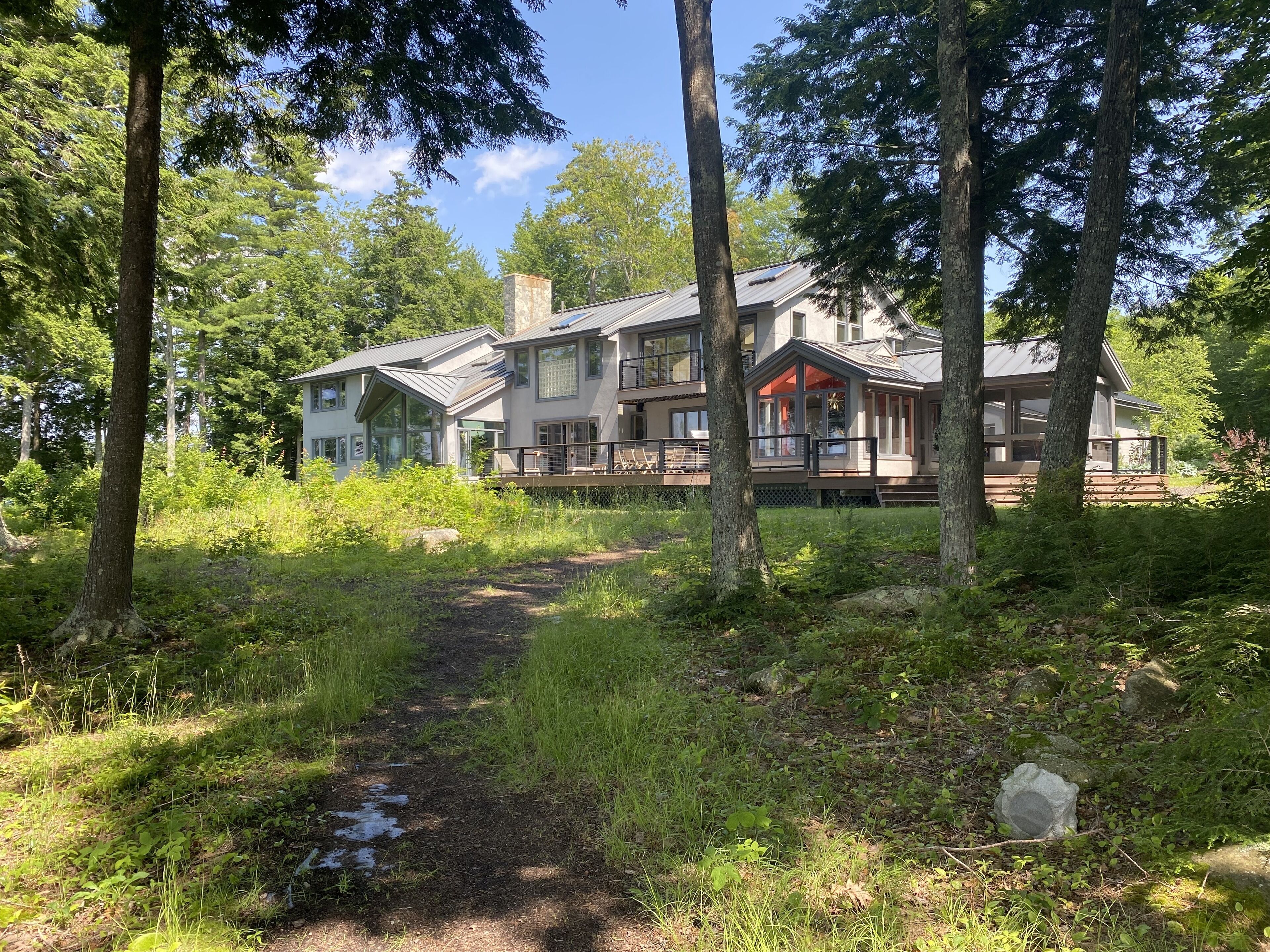 A two-story house with a deck and large windows, surrounded by trees and a grassy path.