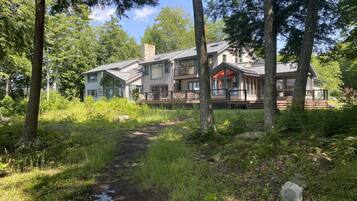 A two-story house with a deck and large windows, surrounded by trees and a grassy path.