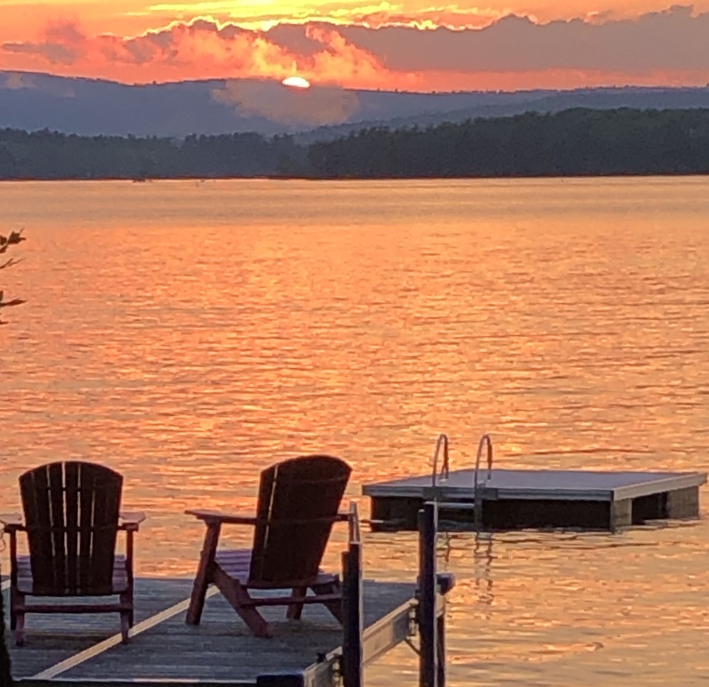A sunset view from a dock with two Adirondack chairs facing a calm lake.