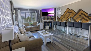 A modern living room with a sofa, coffee table, and a TV mounted on the wall. There is a fireplace with a mantel and a bar area with stools. - SC 5109: Modern 1BR w/private balcony. Perfect for summer with pool! (Snowshoe)