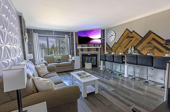 Ein modernes Wohnzimmer mit einer Couch, einem Couchtisch und einem an der Wand montierten Fernseher. Ein Kamin mit Kaminfeuer und eine Bar mit Hockern sind ebenfalls vorhanden.