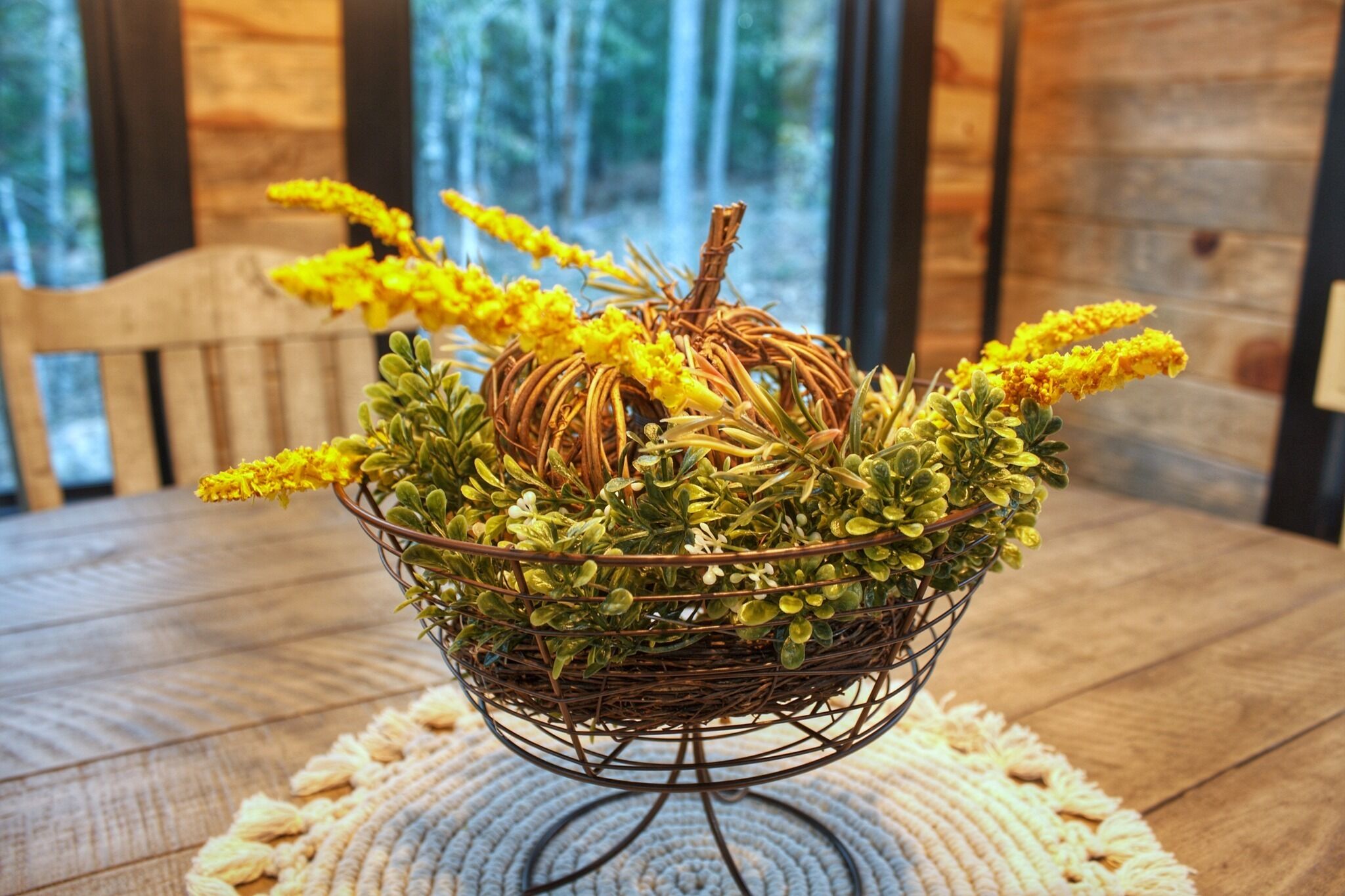 A wicker basket filled with green foliage and yellow flowers, placed on a wooden table with a woven placemat.