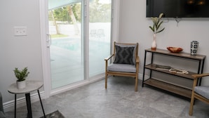 Ein modernes Wohnzimmer mit einer Schiebeglastür, die auf eine Terrasse führt, einem flachbildfernseher an der Wand, einem Holzstuhl mit Kissen, einem kleinen Beistelltisch mit Pflanze und einer Konsole mit dekorativen Gegenständen.
