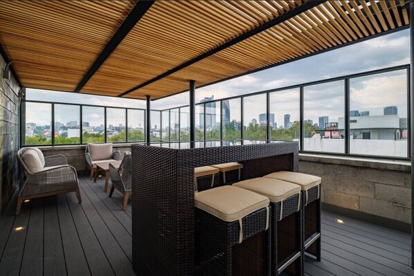 A rooftop terrace with wicker furniture, large windows, and a city view.