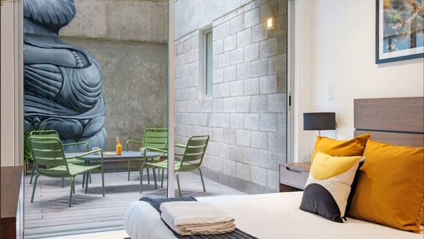 A modern hotel room with a bed, a small table with chairs, a large wall mural, and a window.
