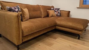 Ein braunes Ecksofa mit floralen Kissen, platziert in einem Raum mit Holzboden und einem Fenster mit Jalousien.