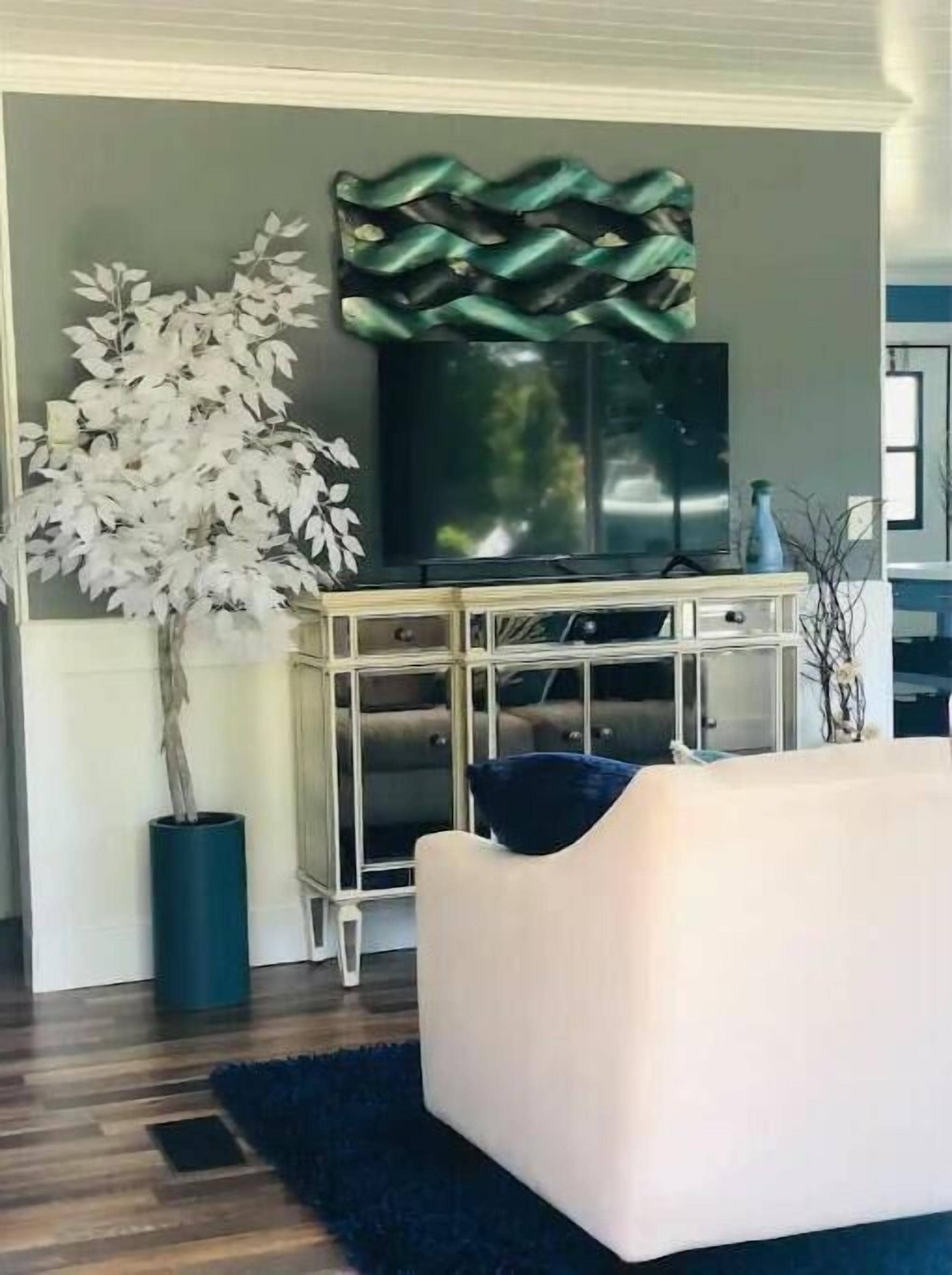 Un salon avec un canapé blanc, une grande plante en pot, un élément décoratif mural et un téléviseur posé sur un meuble.