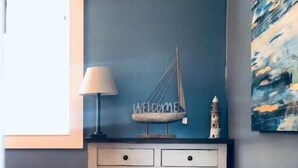 A bedroom with a bed, a bedside table with a lamp, a wall-mounted shelf with a 'WELCOME' sign, a small decorative item, and a painting on the wall. - Beautiful Oceanview Retreat 2 Bedroom Home (Port Hadlock-Irondale)