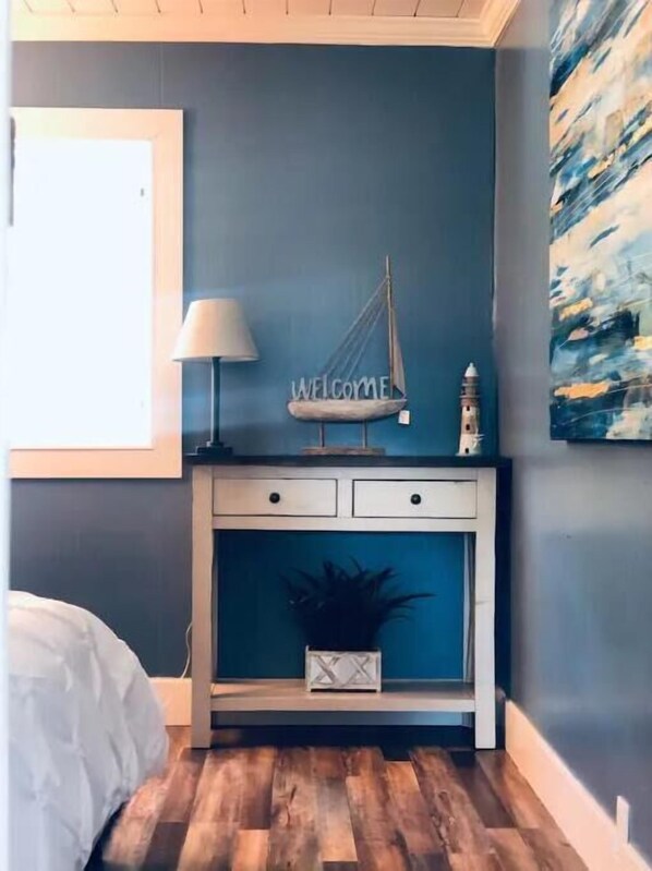 Un dormitorio con cama, una mesita de noche con lámpara, un estante de pared con un cartel que dice 'WELCOME', un adorno pequeño y un cuadro en la pared.