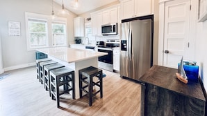 A modern kitchen with white cabinets, stainless steel appliances, a central island with bar stools, and pendant lighting. - OceansideLuxuryCondo*Walk2Beach*Pool*Pets*NoStairs (Corolla)