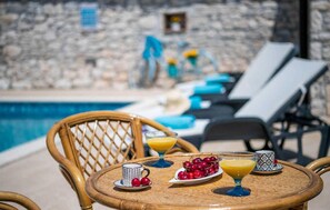 Mesa junto a la piscina con bebidas, fruta, sillas de descanso y un muro de piedra al fondo.