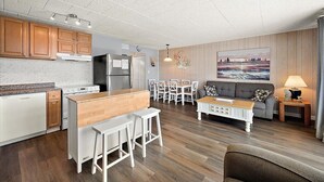 Une cuisine moderne avec des meubles en bois, un coin repas avec des chaises blanches, un canapé et un tableau accroché au mur.