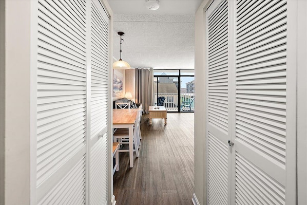 A hallway with a wooden floor, white sliding doors, a dining area with a table and chairs, and a view of buildings through large windows.