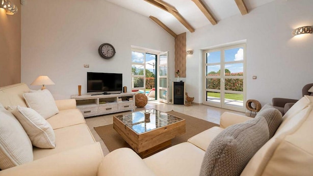 A spacious living room with a large TV, comfortable seating, a fireplace, and large windows offering a view of the outdoors.