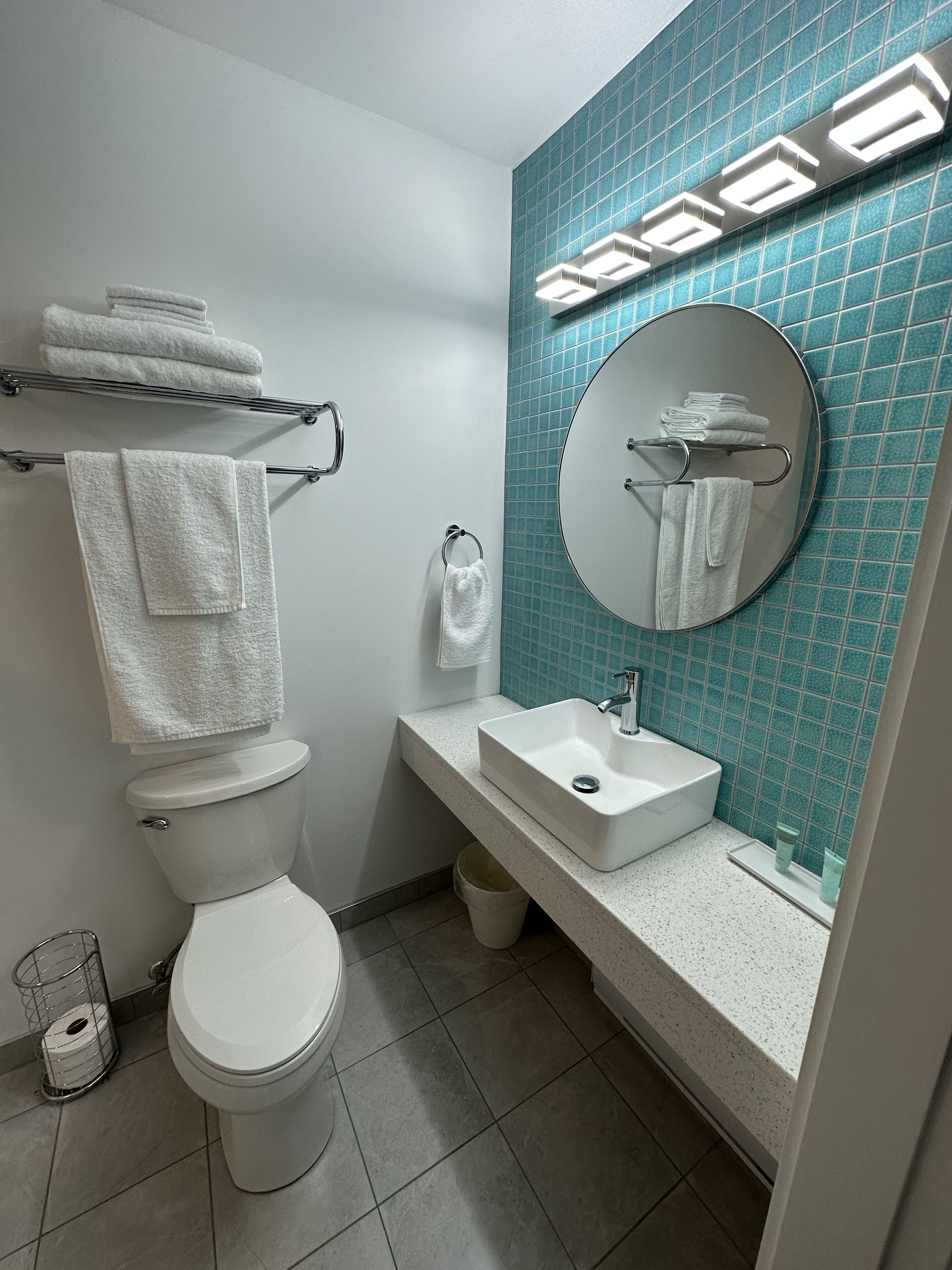 Een moderne badkamer met toilet, wastafel en spiegel. Handdoeken liggen op de handdoekhouder en er staat een prullenbak.