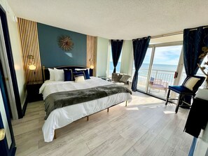 A spacious bedroom with a large bed, a seating area with a chair, and a view of the sea through the windows.