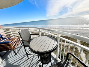A deck with a round table and chairs, overlooking a beach.