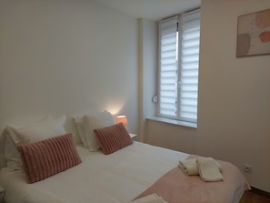 Un dormitorio con cama, almohadas, una lámpara de noche, una ventana con persianas y una obra de arte colgada en la pared.