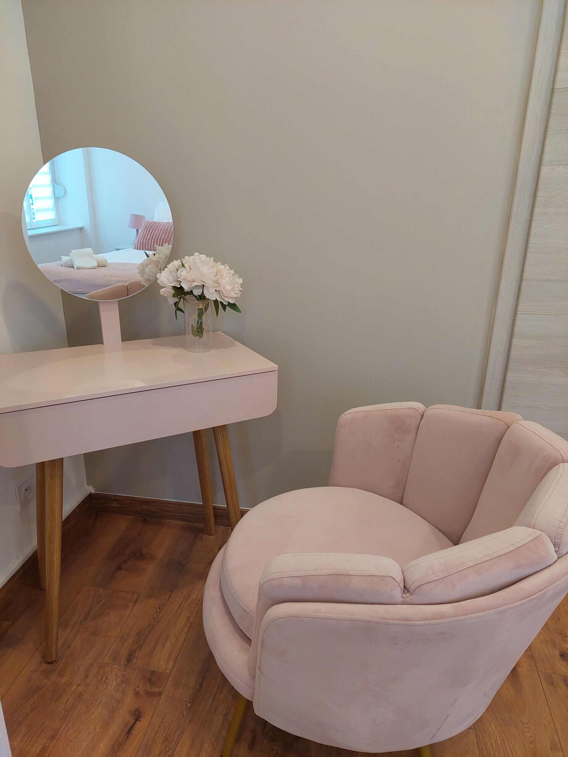 Una habitació amb una taula de tocador, un mirall rodó, un vas amb flors i un butac armchair confortable.