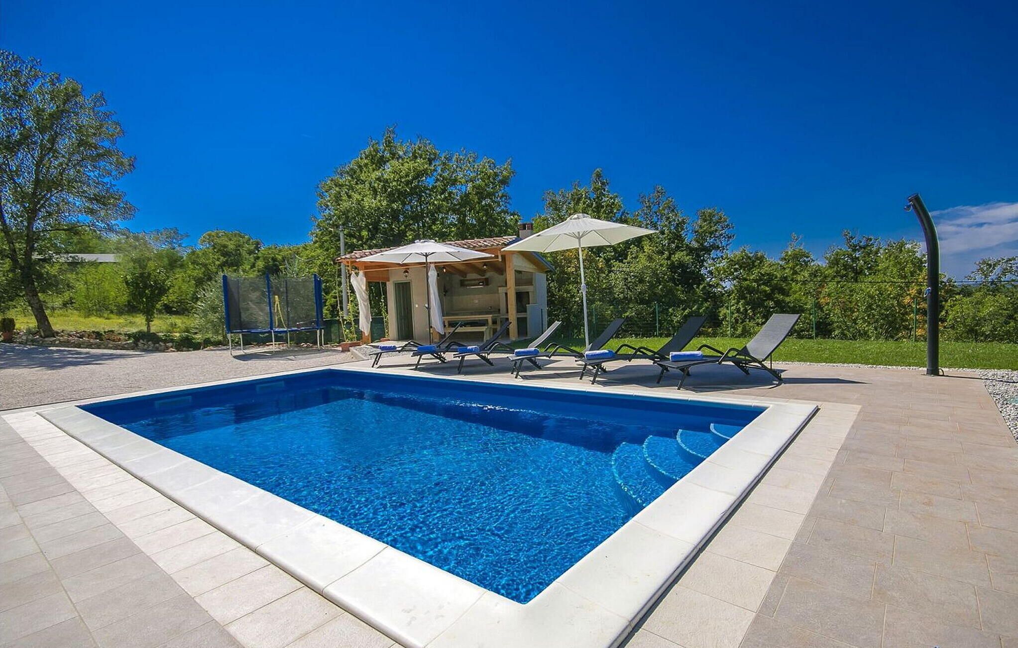 Una piscina rettangolare con bordo piastrellato, circondata da un solarium lastricato. Si vedono lettini da spiaggia e un piccolo gazebo con tetto e ombrellone.