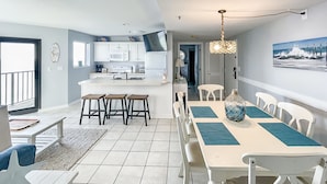 Une cuisine moderne avec des meubles blancs, un îlot central doté de tabourets, un coin repas avec une longue table et des chaises, et une vue sur l’océan par une fenêtre.