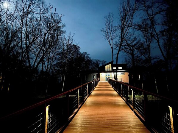 A wooden bridge leading to a building with a modern design, surrounded by trees.