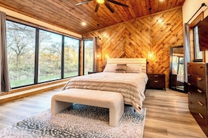 A bedroom with a large bed, a bench, a dresser, and a mirror. There are windows providing a view of trees and a body of water.