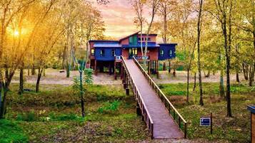 A two-story wooden house with a deck leading to the entrance, surrounded by trees and a sunset in the background.