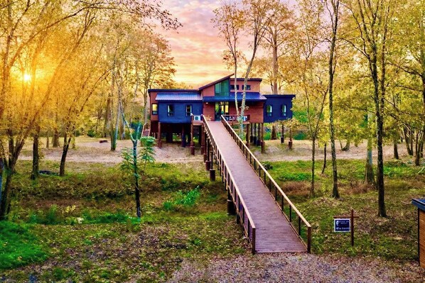 A two-story wooden house with a deck leading to the entrance, surrounded by trees and a sunset in the background. - All is Found Riverfront Cabin with Sauna (Broken Bow)