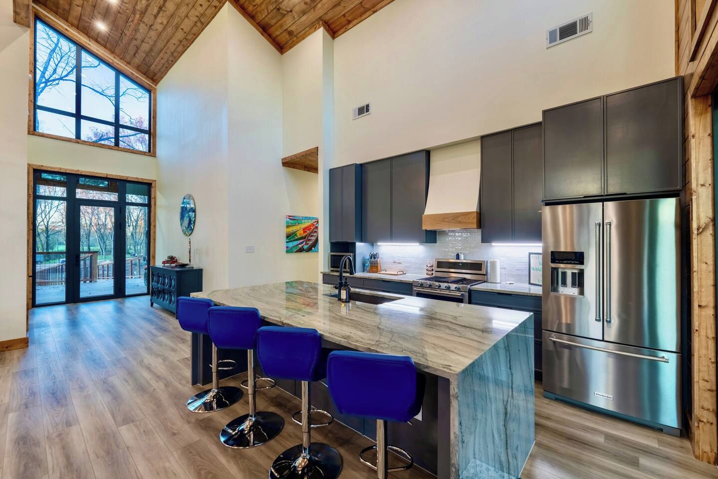 A modern kitchen with a large island, bar stools, stainless steel appliances, and a view through the glass doors.