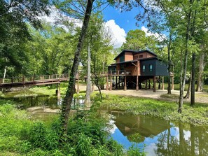 A house with a deck, elevated on stilts, surrounded by water and greenery.
