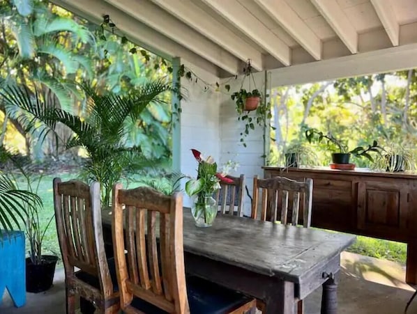 A covered outdoor dining area with a wooden table and chairs, surrounded by potted plants and a view of greenery.