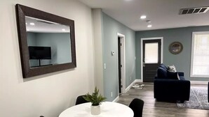Ein Essbereich mit einem runden Tisch, schwarzen Stühlen und einer Zimmerpflanze. Im Hintergrund befindet sich eine dunkel gefärbte Couch und ein kleiner Tisch.