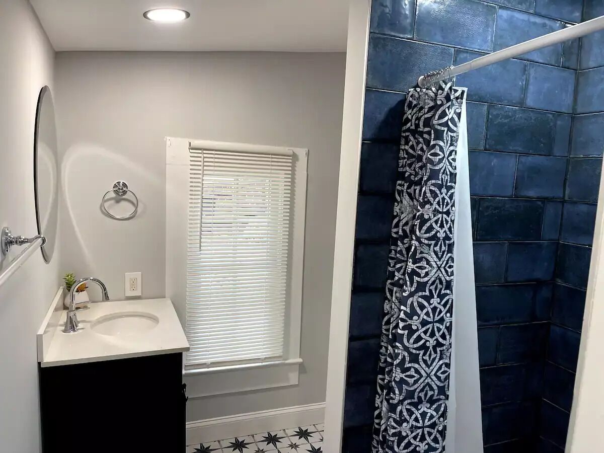A bathroom with a white sink, a mirror, a window with blinds, and a shower curtain with a geometric pattern.