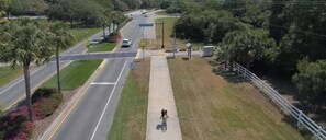 A road with a cyclist on a sidewalk, surrounded by greenery and a white fence.