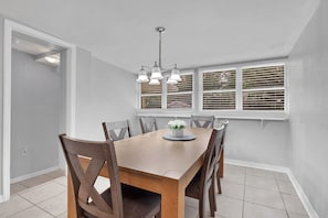 A dining area with a wooden table, six chairs, a chandelier, and windows with blinds. - Home w/ Fenced Yard, Hot tub, Grill & 3 King Beds! (Sarasota)
